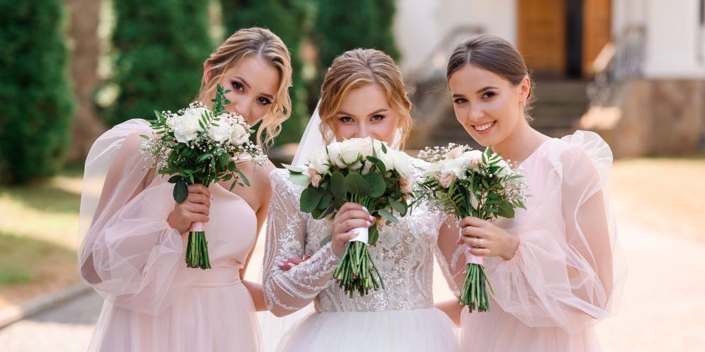 Portrait of elegant bridesmaids and bride in white puffy dress, holding bouquets with flowers waiting for wedding ceremony while posing outdoors.